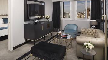 Habitación de hotel moderna con cama, sofá, televisión, una mesita con un jarrón de flores y vistas a la ciudad desde la ventana.