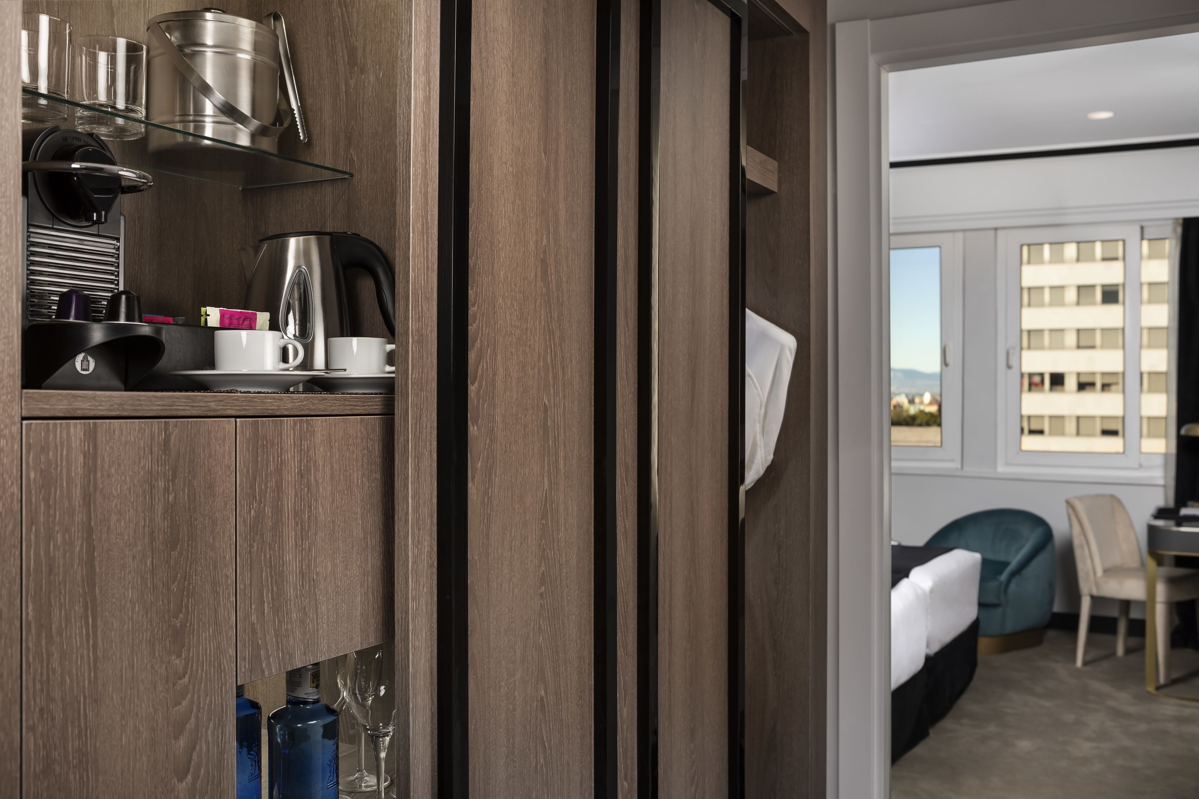 Habitación de hotel con un armario de madera que contiene utensilios de cocina, una cama con edredón blanco, una silla color turquesa y vistas a un edificio por la ventana.