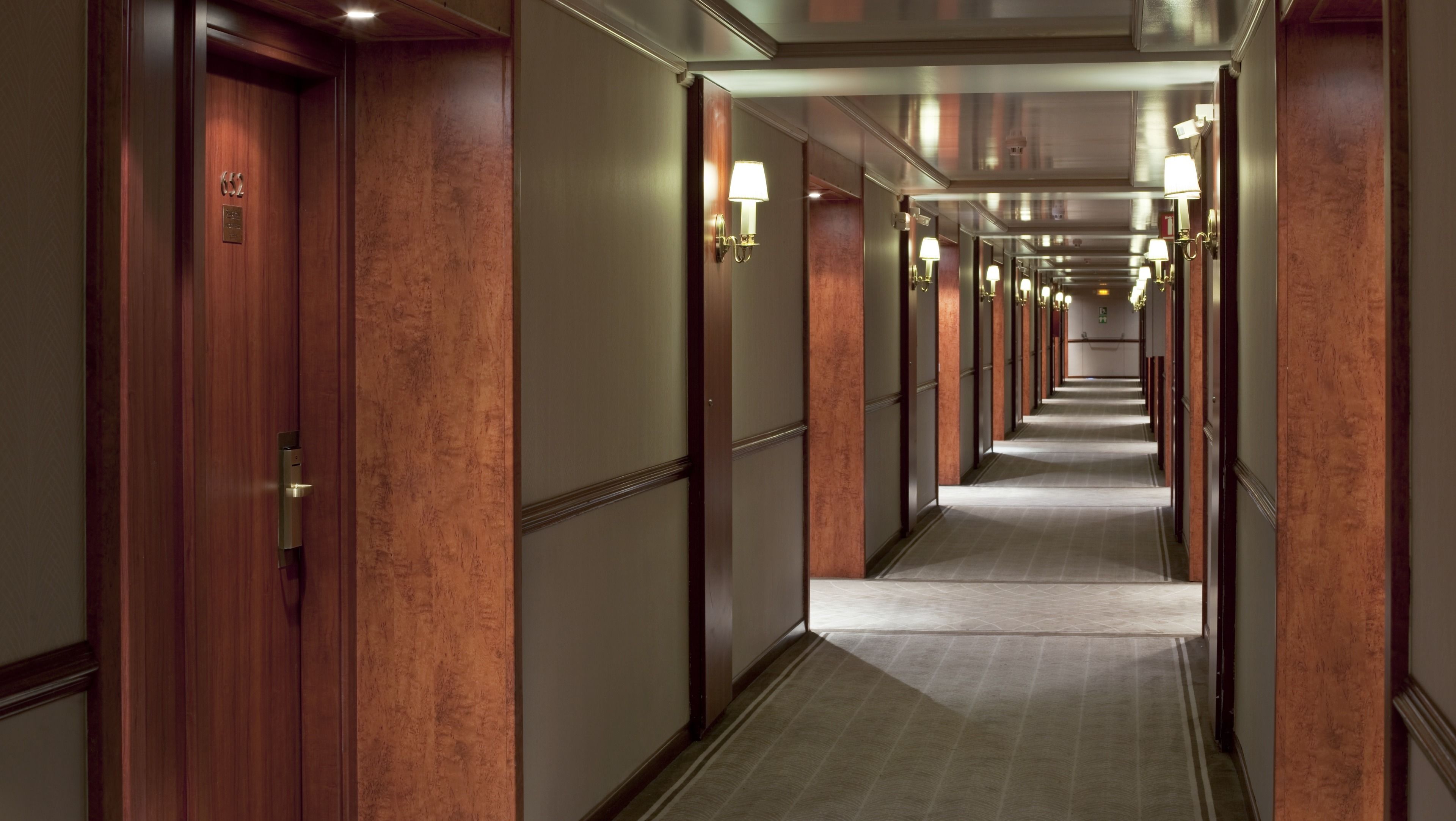 A hotel corridor with wooden doors, a carpeted floor, and wall-mounted lamps.