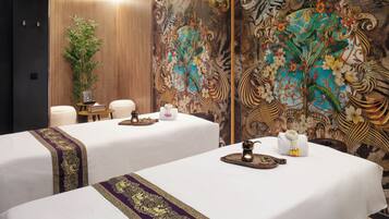 A room with two beds, each covered with white linens and purple decorative borders. There is a small table with a tea set and a potted plant. The walls are adorned with intricate floral and scroll designs.