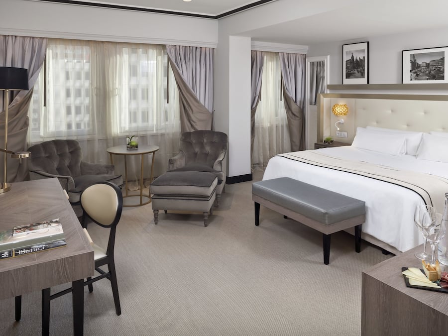 Une chambre d’hôtel avec un grand lit, deux fauteuils, une petite table et un bureau avec une lampe.