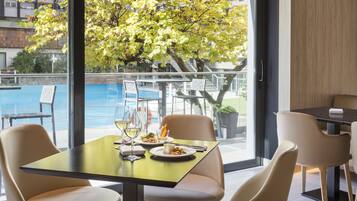 A dining area with a table set for two, a view of a swimming pool, and a tree outside the window.