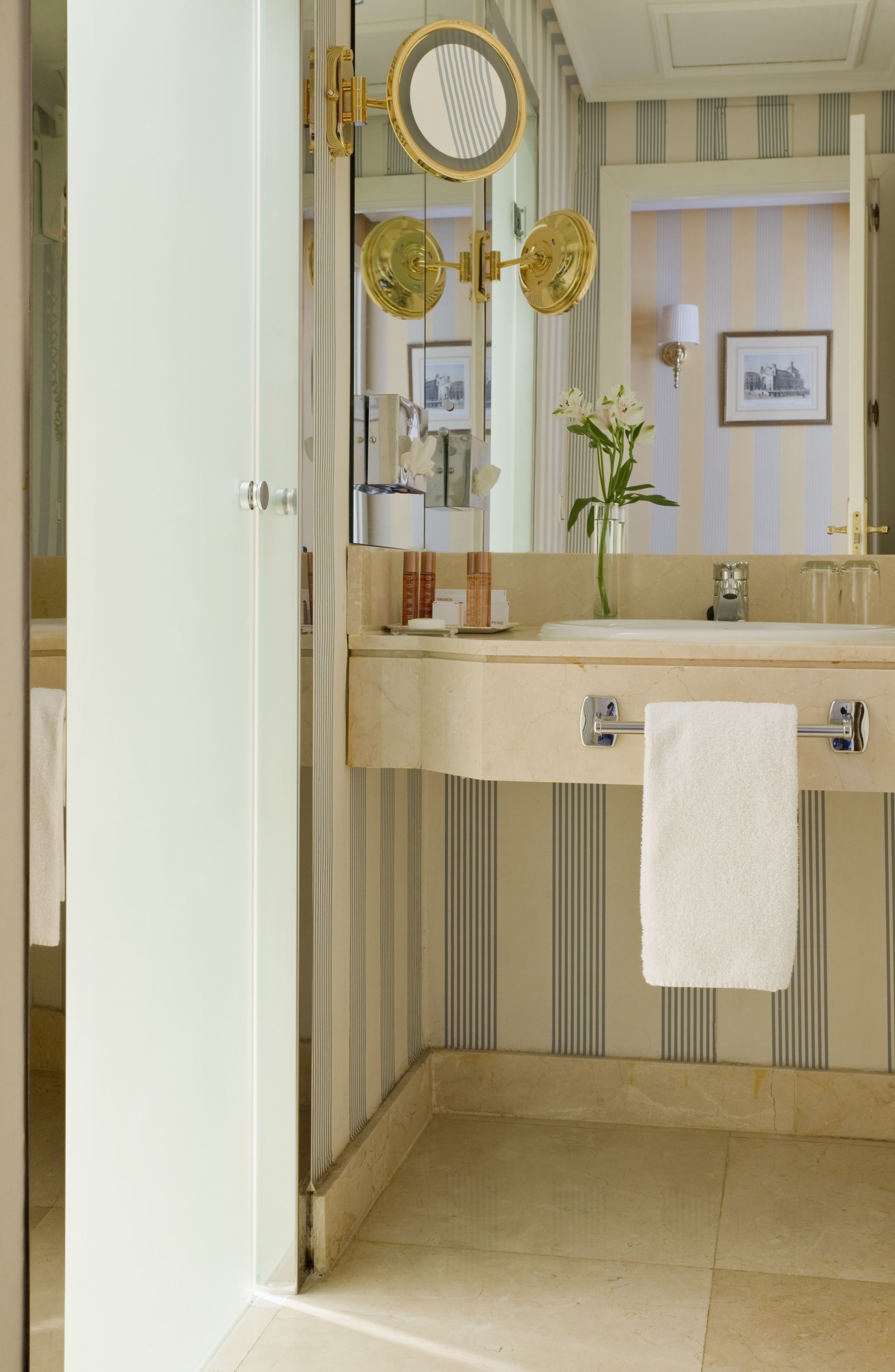 Ein Badezimmer mit Waschtisch, einem Spiegel mit goldener Rahmung, einem an der Wand hängenden Handtuch und gestreifter Tapete.