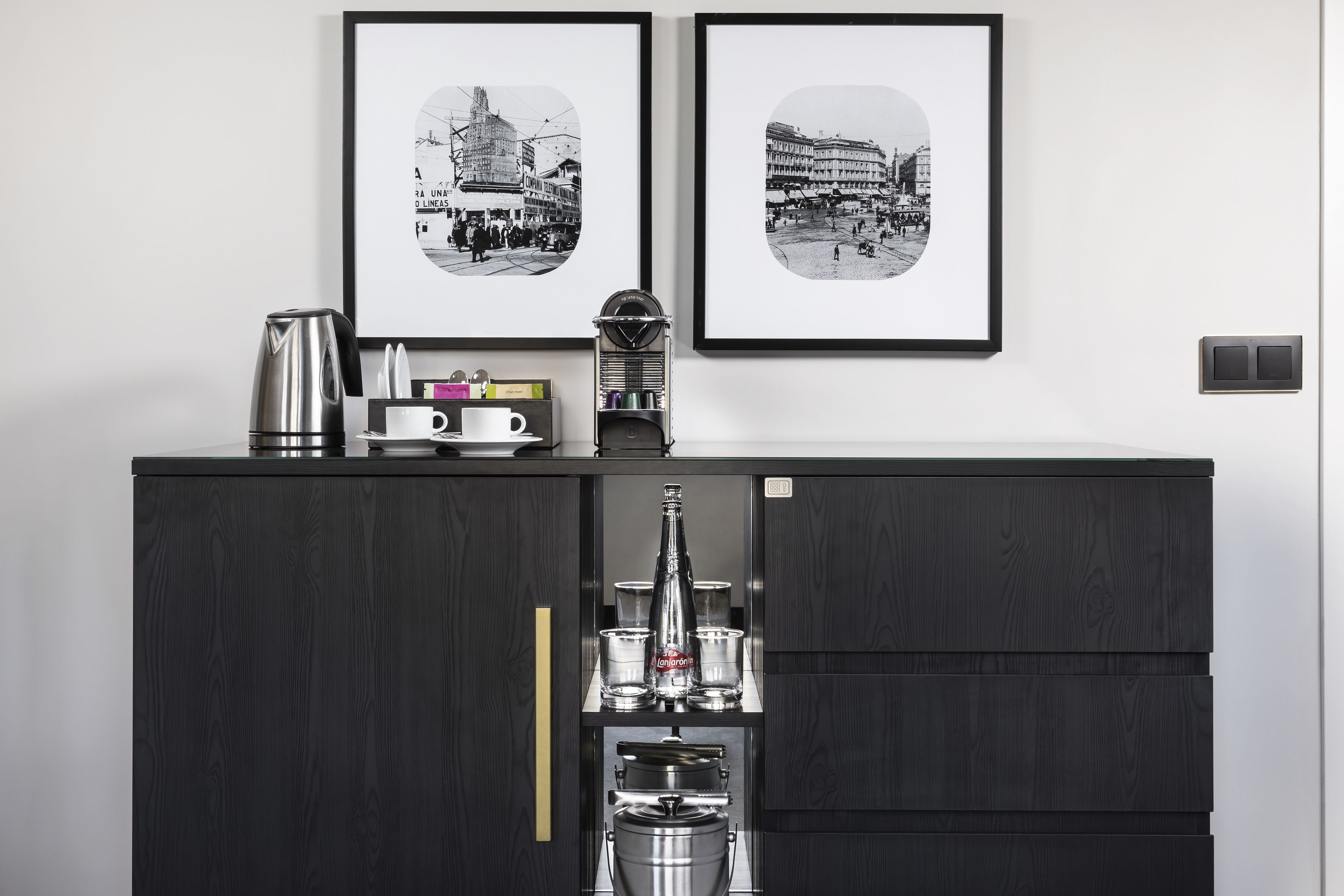 A kitchen area with a dark wood cabinet, a coffee maker, and a kettle. There are two framed black and white photographs on the wall above the cabinet.