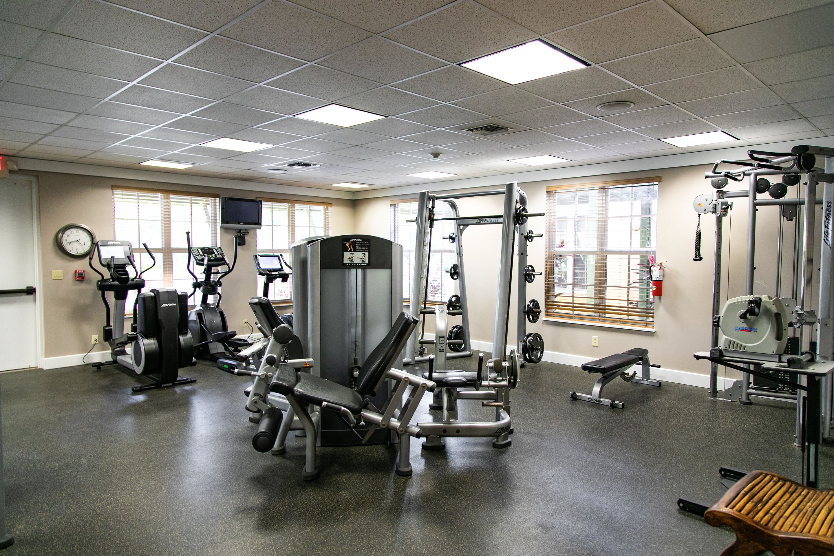 A gym with various exercise equipment, a clock on the wall, and windows with blinds.