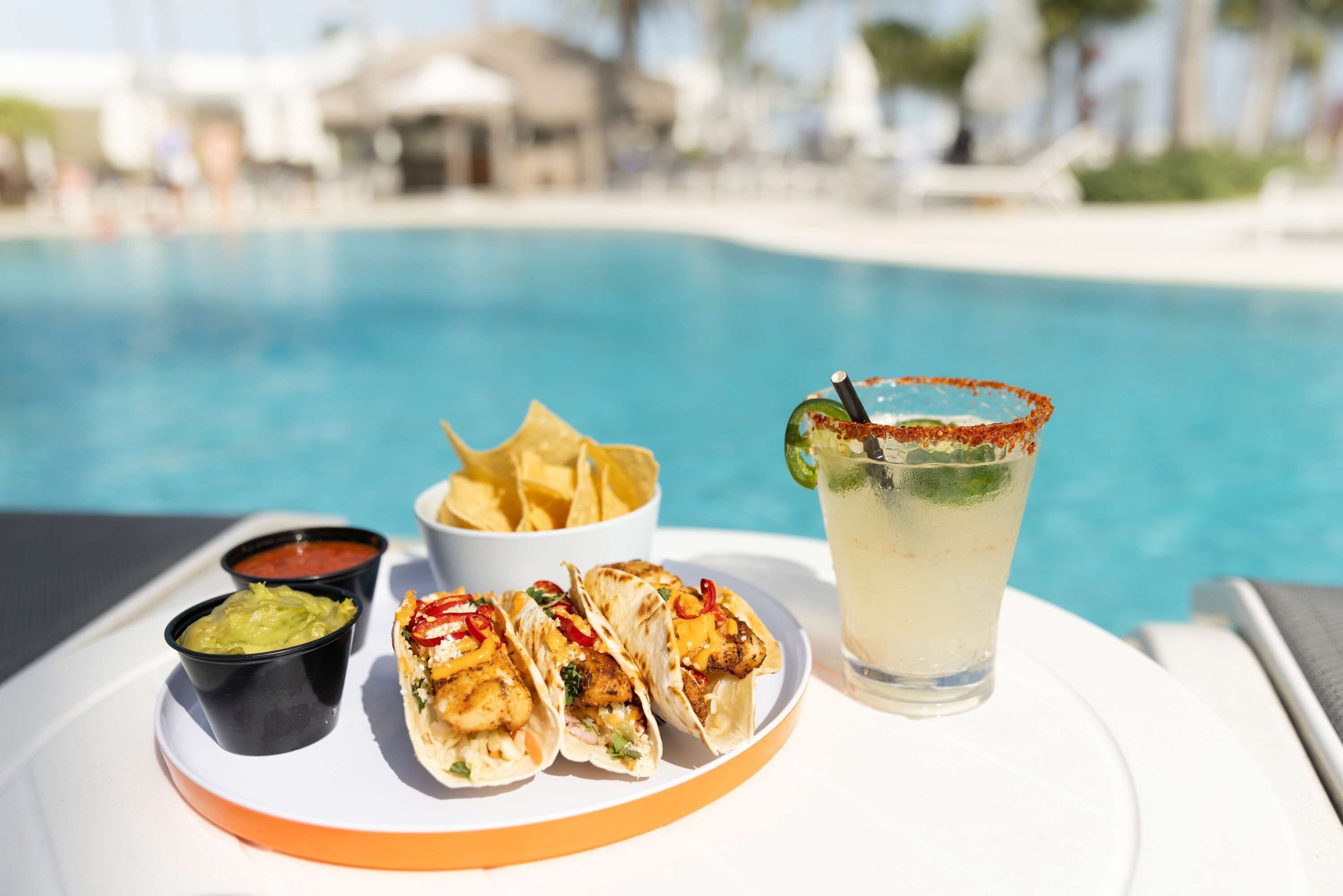 Un plato con tacos, guacamole y una margarita junto a una piscina.