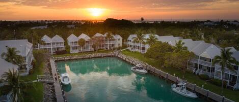 A resort with a central pool, connected by walkways to multiple bungalow-style buildings, surrounded by palm trees and a sunset view.