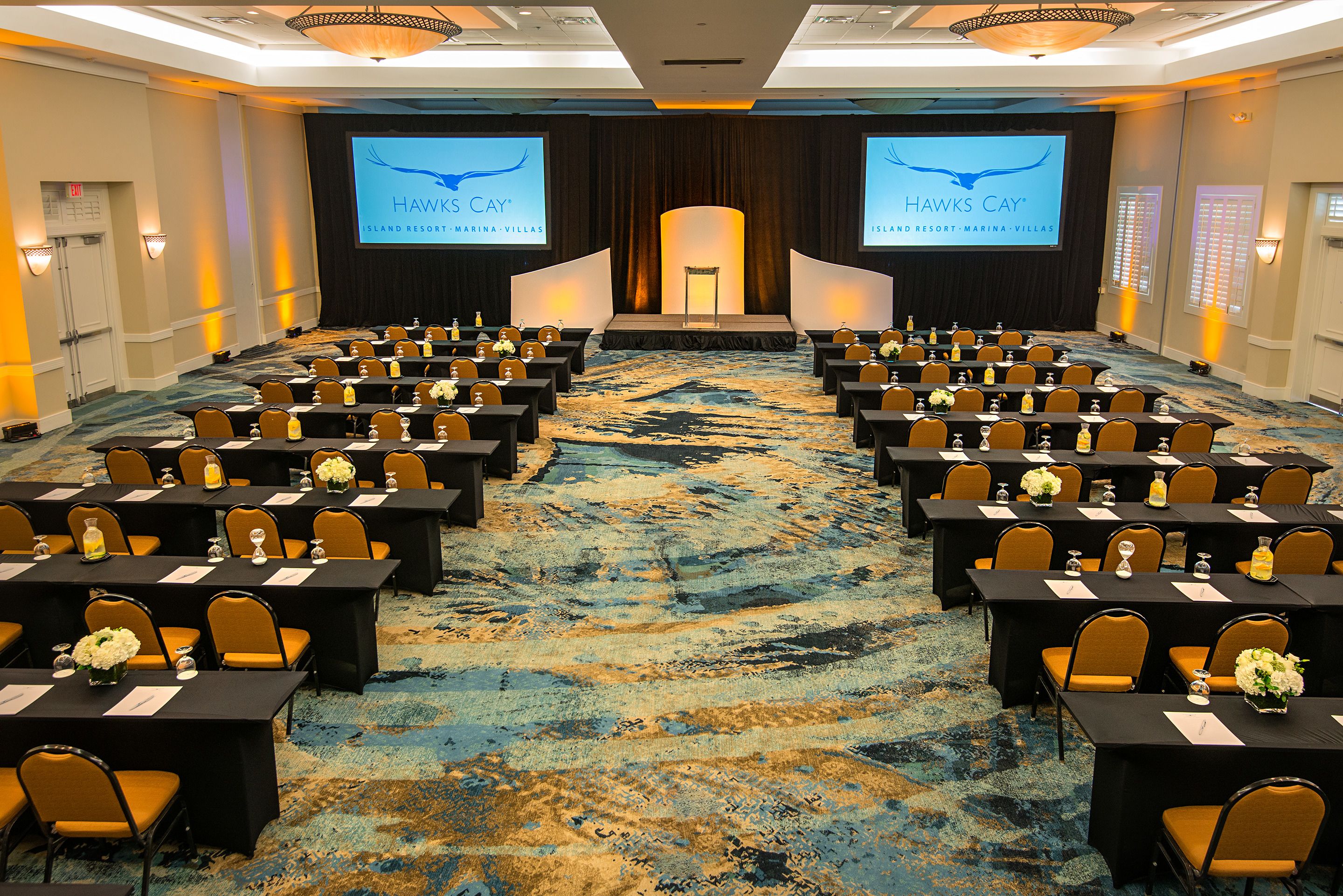 A conference room with rows of tables and chairs, a stage with a screen displaying 'HAWKS CAY' branding, and a carpeted floor.