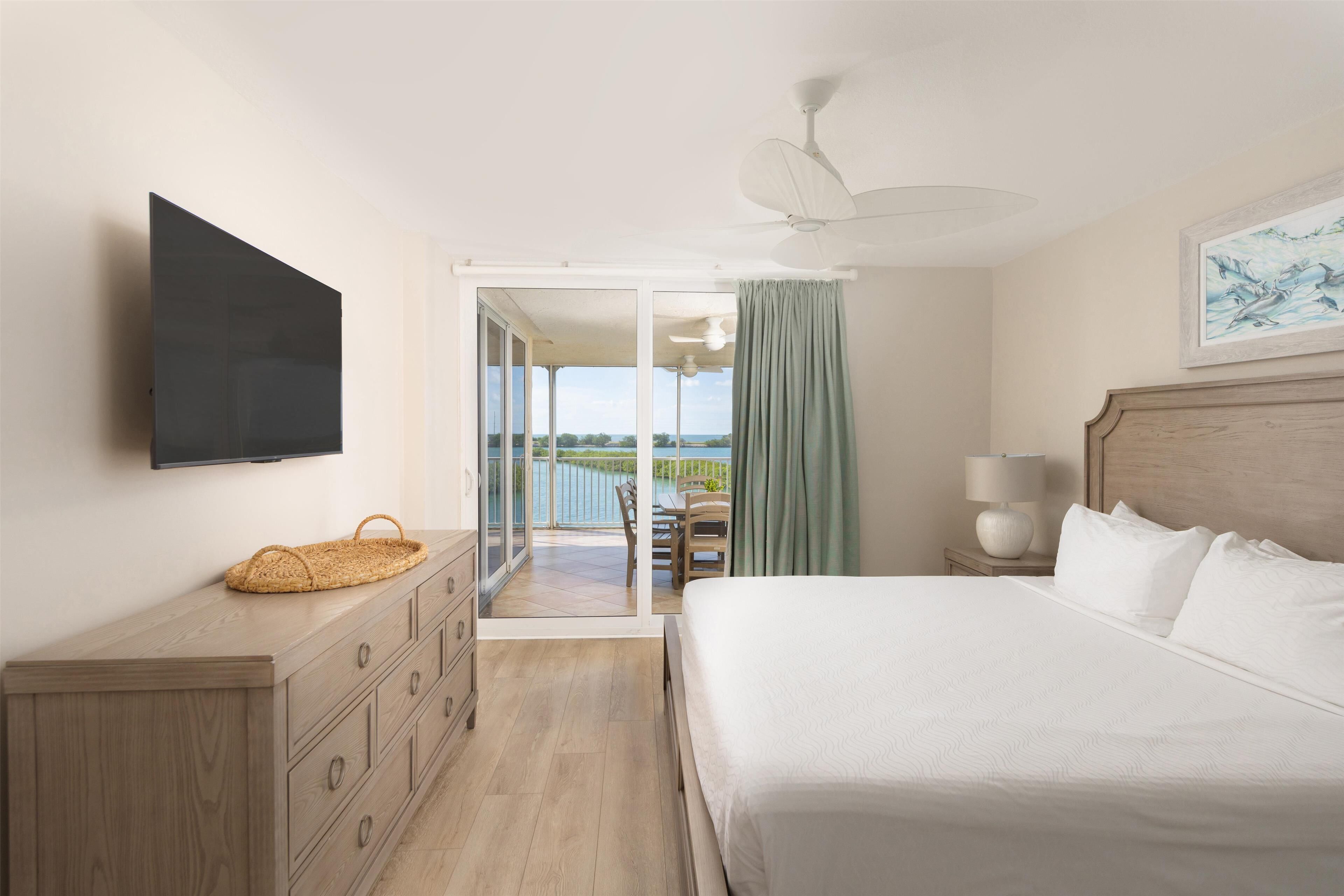 A bedroom with a bed, a TV mounted on the wall, a wooden dresser, a nightstand with a lamp, and a view of the outdoors through a sliding door.