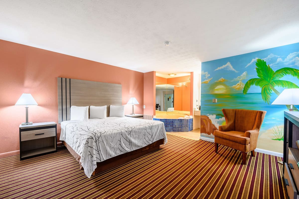 Une chambre d’hôtel avec un lit, deux tables de chevet avec des lampes, une chaise, une grande fresque représentant une scène de plage, et une salle de bain visible à travers une porte ouverte.