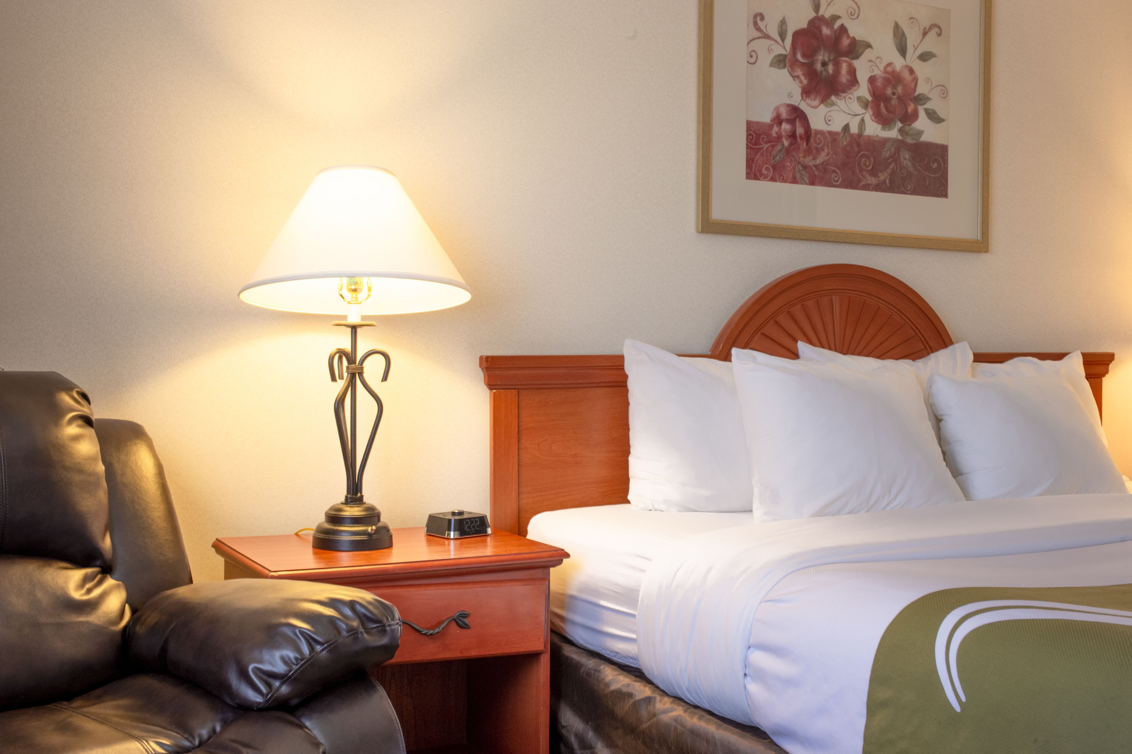 A hotel room with a bed, a nightstand with a lamp, a leather chair, and a framed floral artwork on the wall.