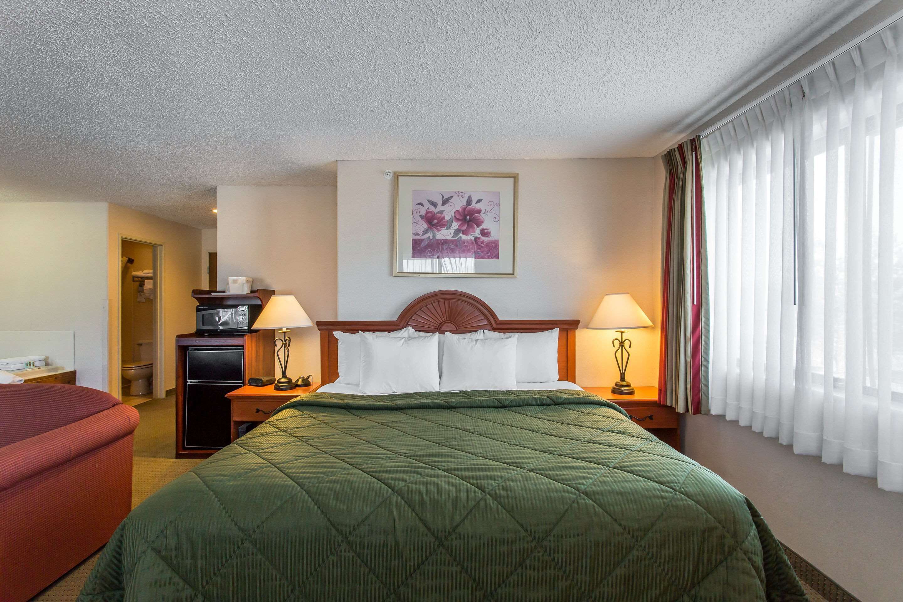Ein Hotelzimmer mit einem Bett, zwei Nachttischlampen, einem gerahmten Blumenbild an der Wand, einem kleinen Schreibtisch mit Lampe und einem roten Sessel.