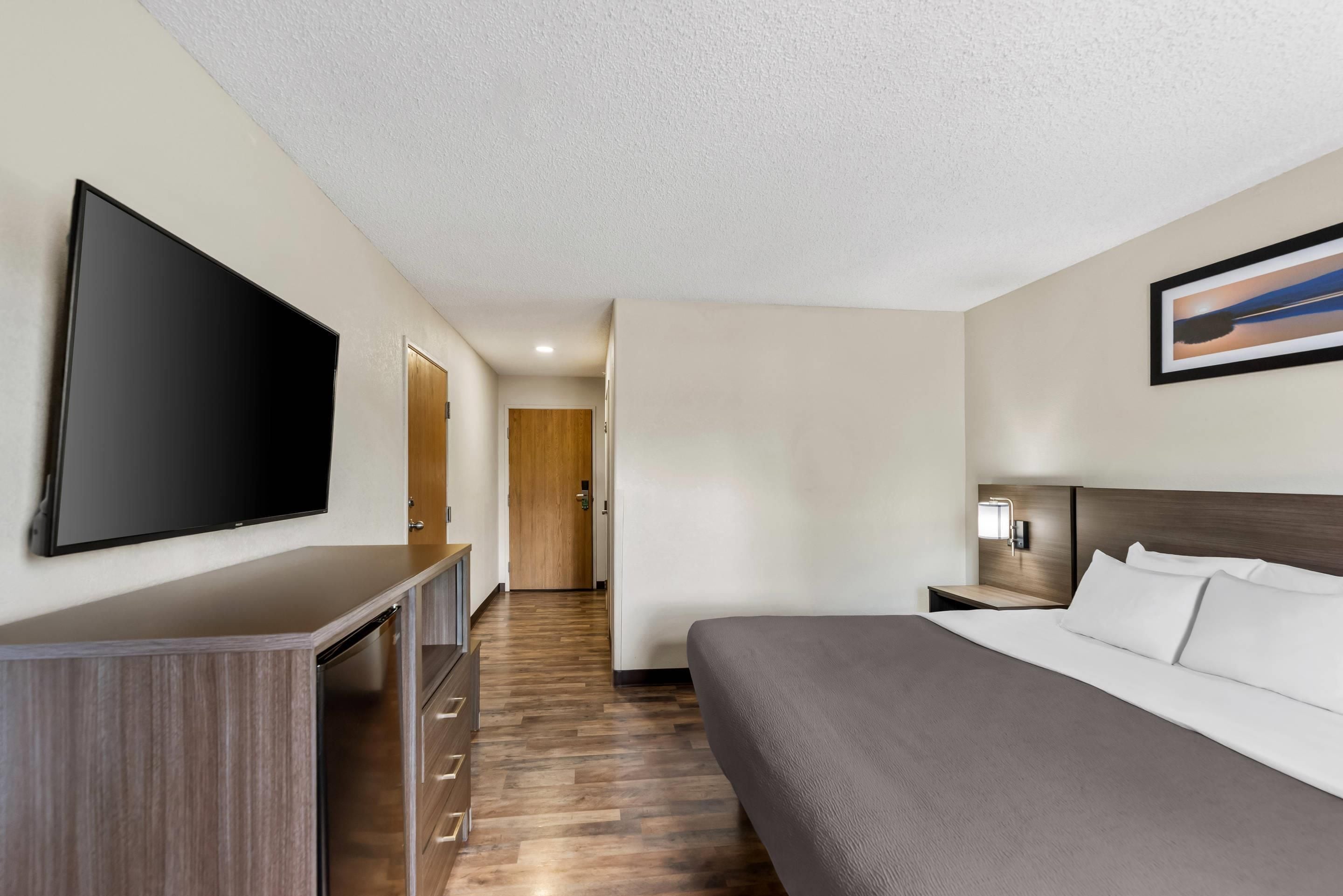 Habitación de hotel con una cama grande, un televisor de pantalla plana montado en la pared, un aparador de madera, una mesita de noche con lámpara y una puerta que conduce a otra habitación.