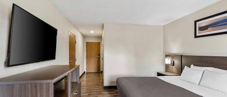 Habitación de hotel con una cama grande, un televisor de pantalla plana montado en la pared, un aparador de madera, una mesita de noche con lámpara y una puerta que conduce a otra habitación.