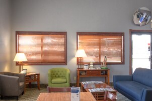 Una sala de estar con dos ventanas, un sillón verde, un sofá azul, una mesa de madera con revistas y una lámpara.