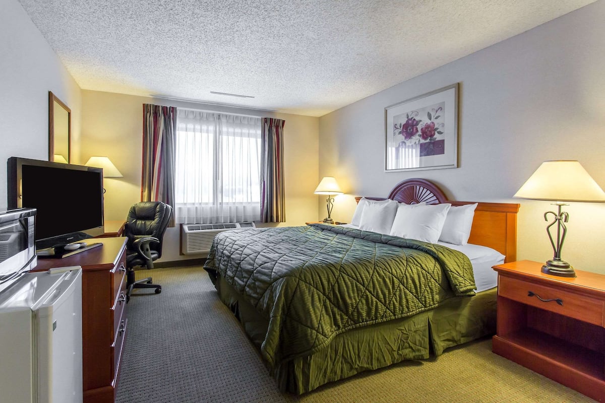 A hotel room with a bed, a desk with a computer, a chair, a TV, and a window with curtains.