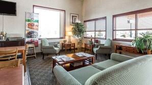 A cozy living room with a sofa, chairs, a coffee table, and a dining area with a table and chairs. There is a TV mounted on the wall, a window with blinds, and a plant on the windowsill.