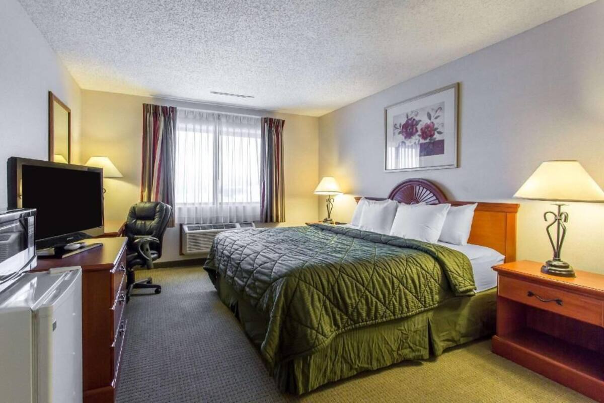 A hotel room with a bed, a desk with a computer, a chair, a TV, and a window with curtains.