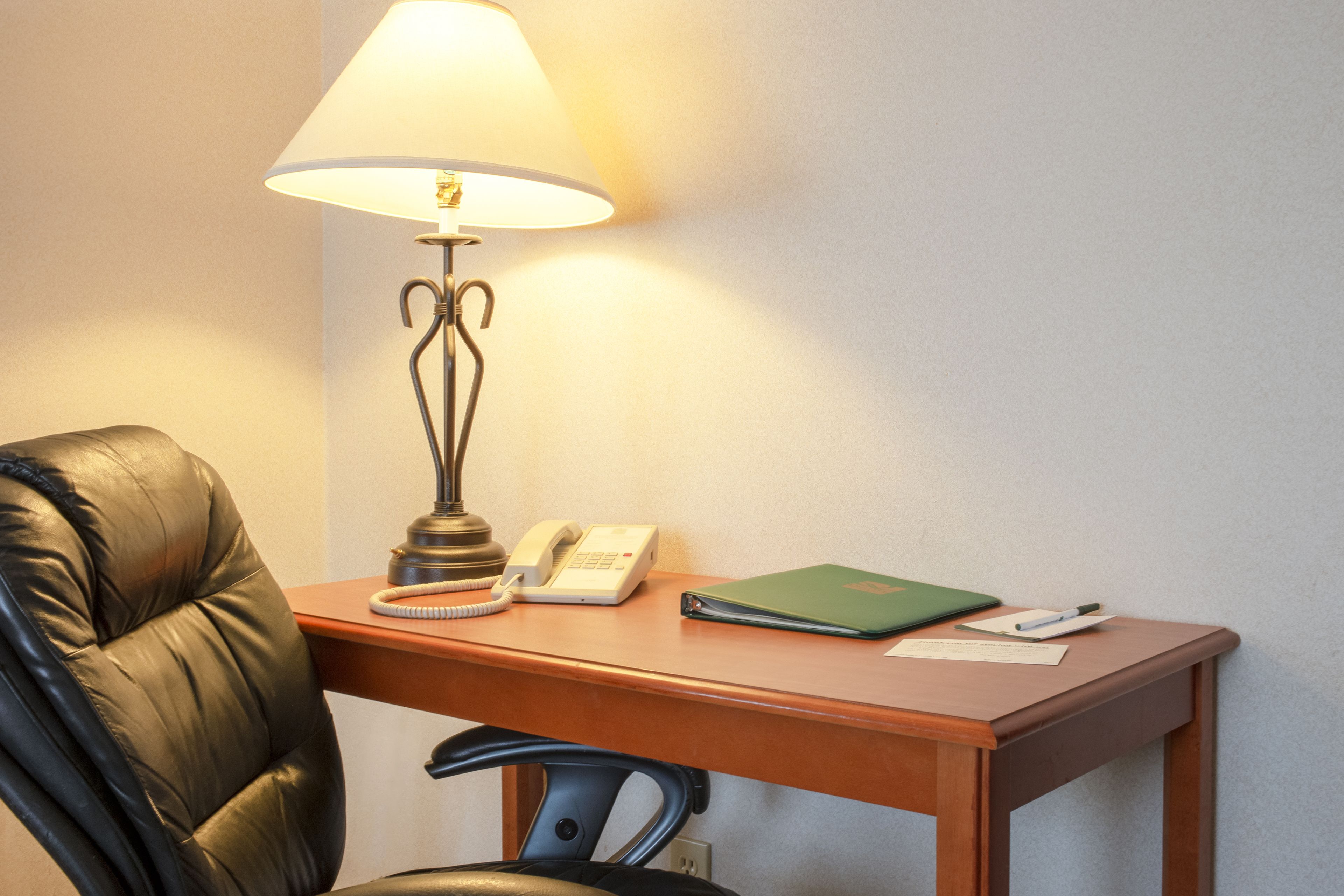 A hotel room with a wooden desk, a leather chair, a lamp, a telephone, a green folder, and a pen.