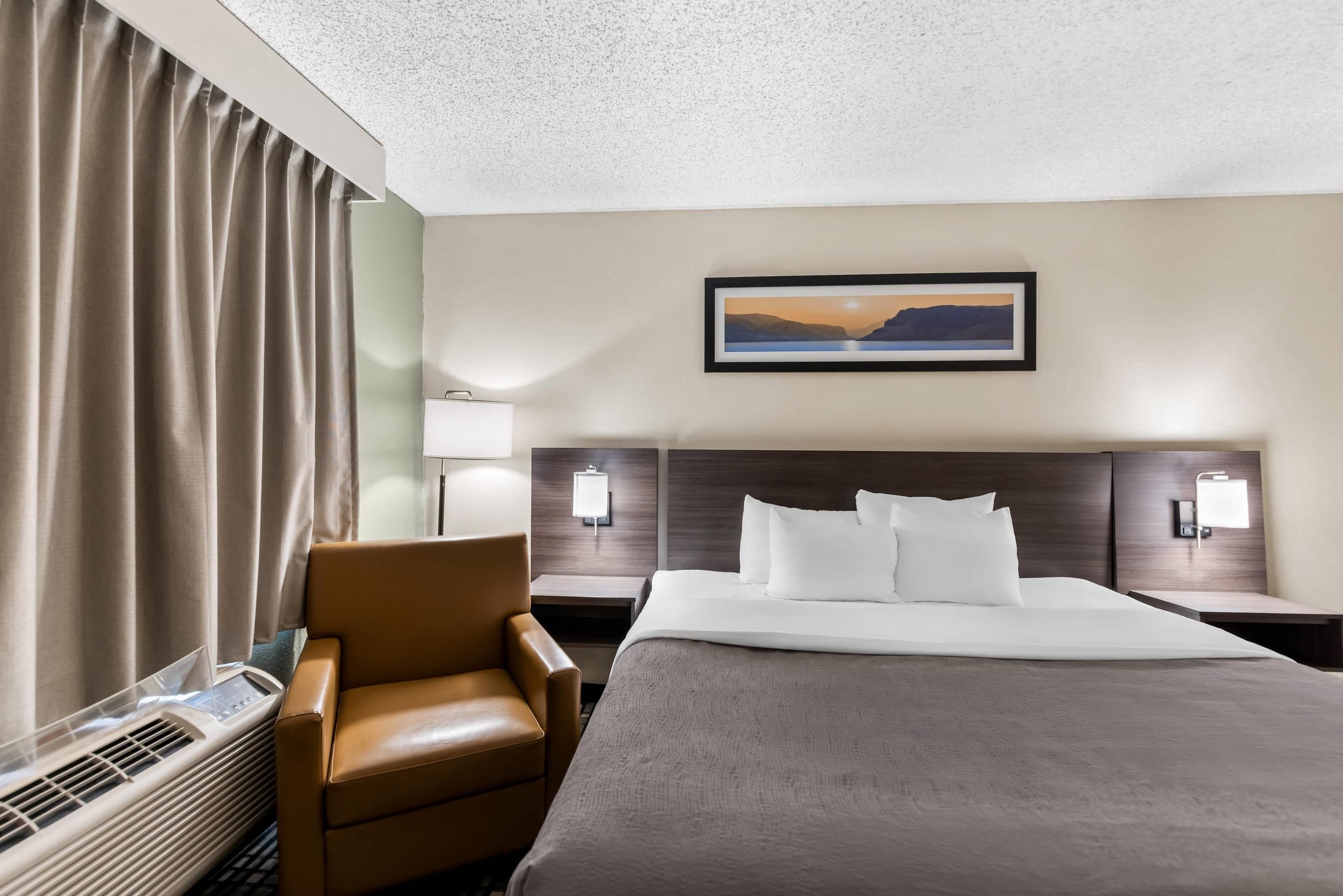A hotel room with a bed, a chair, a desk, a lamp, and a painting on the wall.
