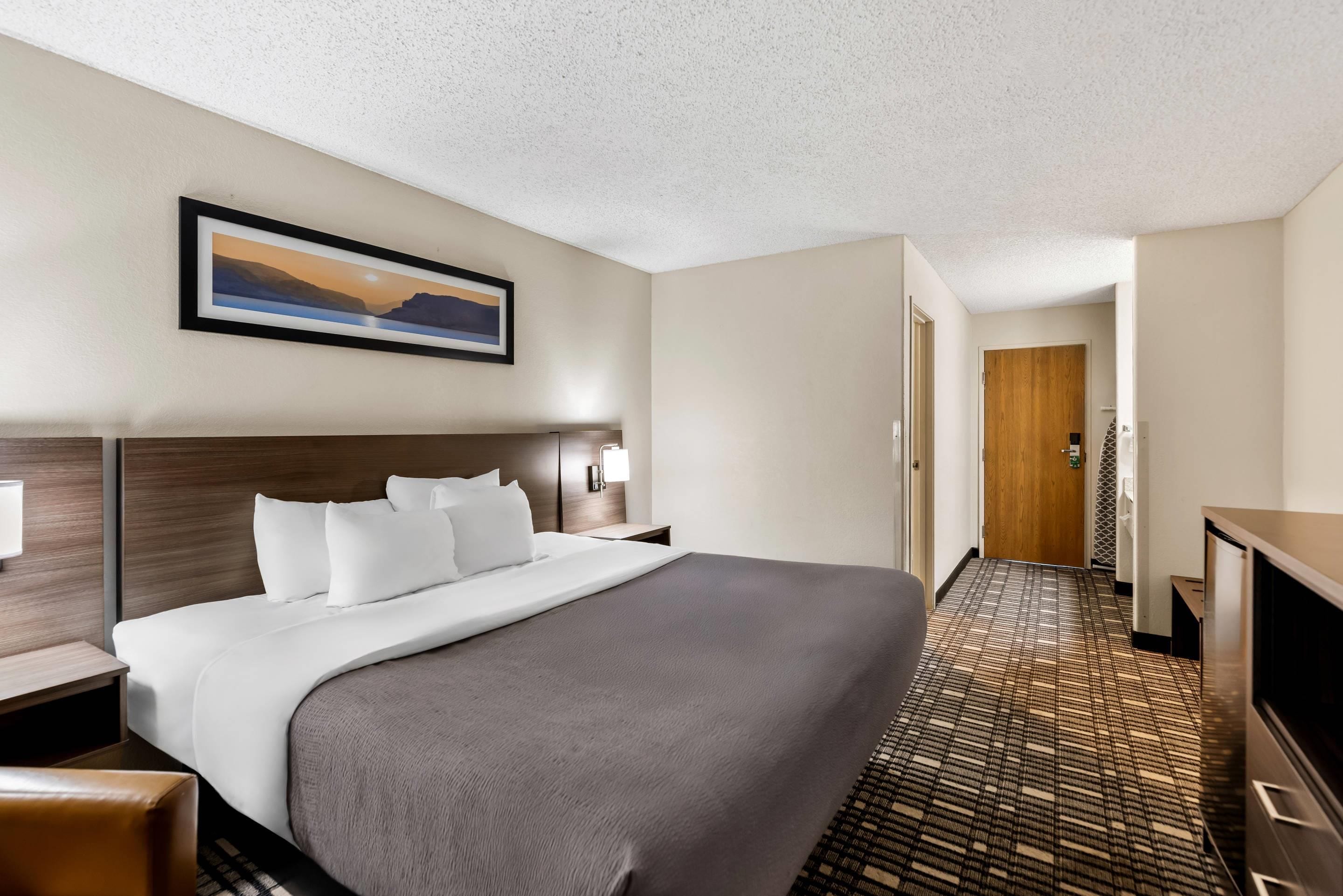 A hotel room with a large bed, a headboard, a bedside table, a lamp, a television, a carpeted floor, and a door leading to another room.