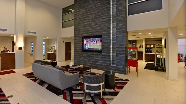 A spacious lobby with a modern design, featuring a large flat-screen TV mounted on a textured wall, a reception desk, and a seating area with a sofa and armchairs.