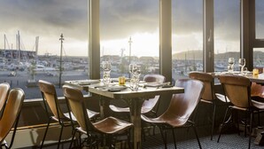 A restaurant with tables set for dining, large windows offering a view of a marina and cityscape.