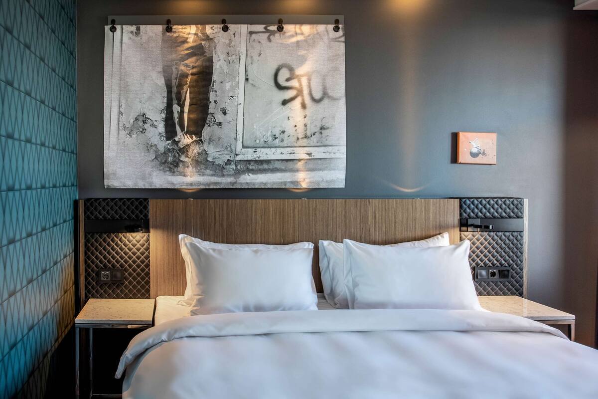 Una habitación de hotel moderna con una cama grande, un cabecero de diseño texturizado, una obra de arte colgada en la pared y una mesita de noche pequeña.