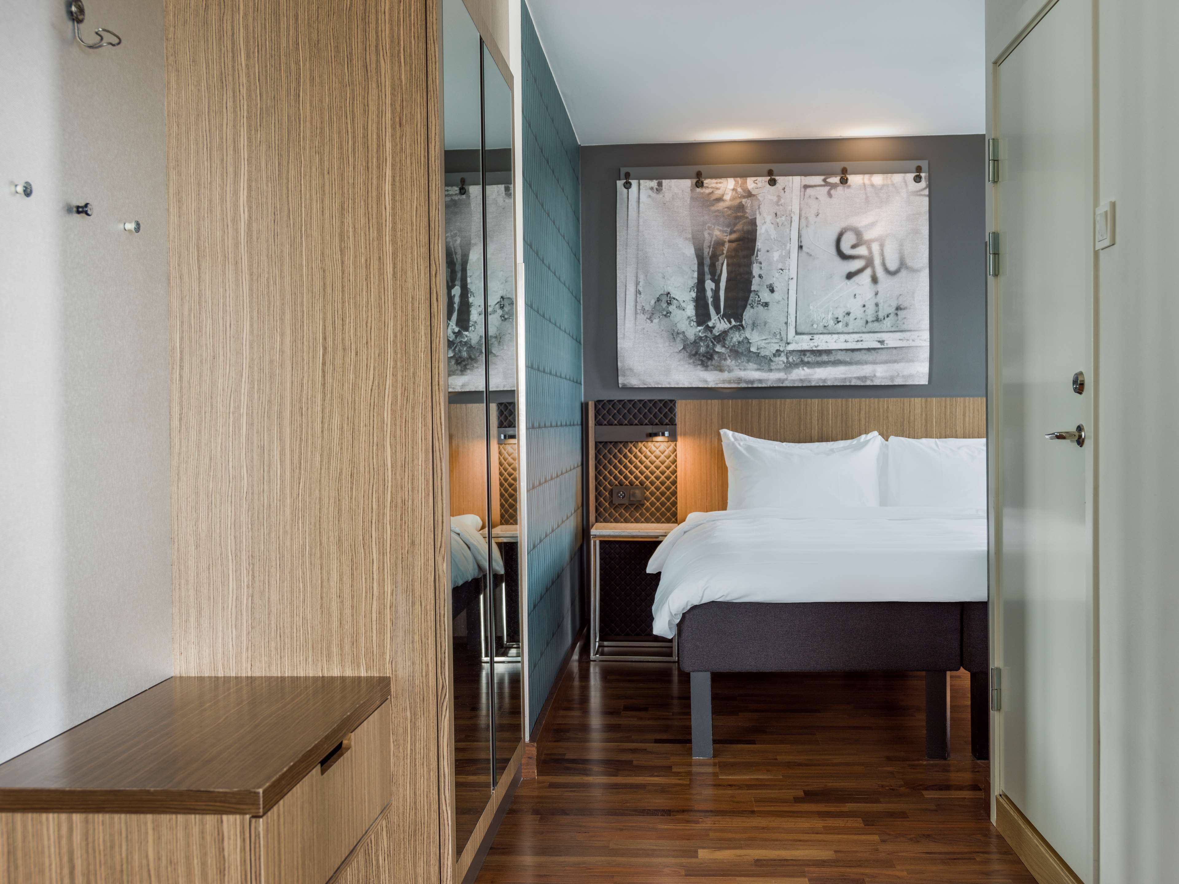 A modern hotel room with a wooden floor, a bed with white linens, a large abstract painting on the wall, and a wooden wardrobe.