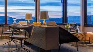 A modern dining area with a central table, chairs, and lamps. Large windows provide a view of the mountains.