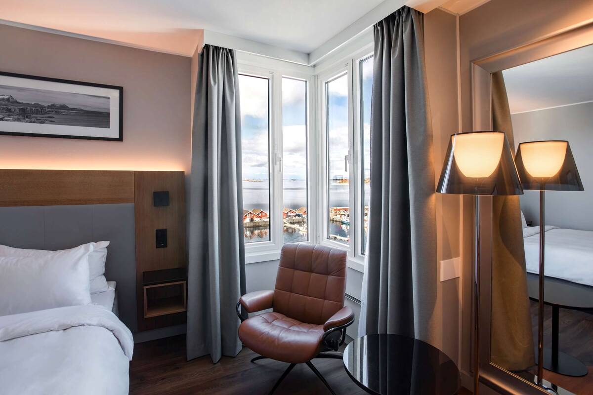 Una habitación de hotel con una cama, un sillón de cuero marrón, una mesita, una lámpara y ventanas grandes con cortinas.
