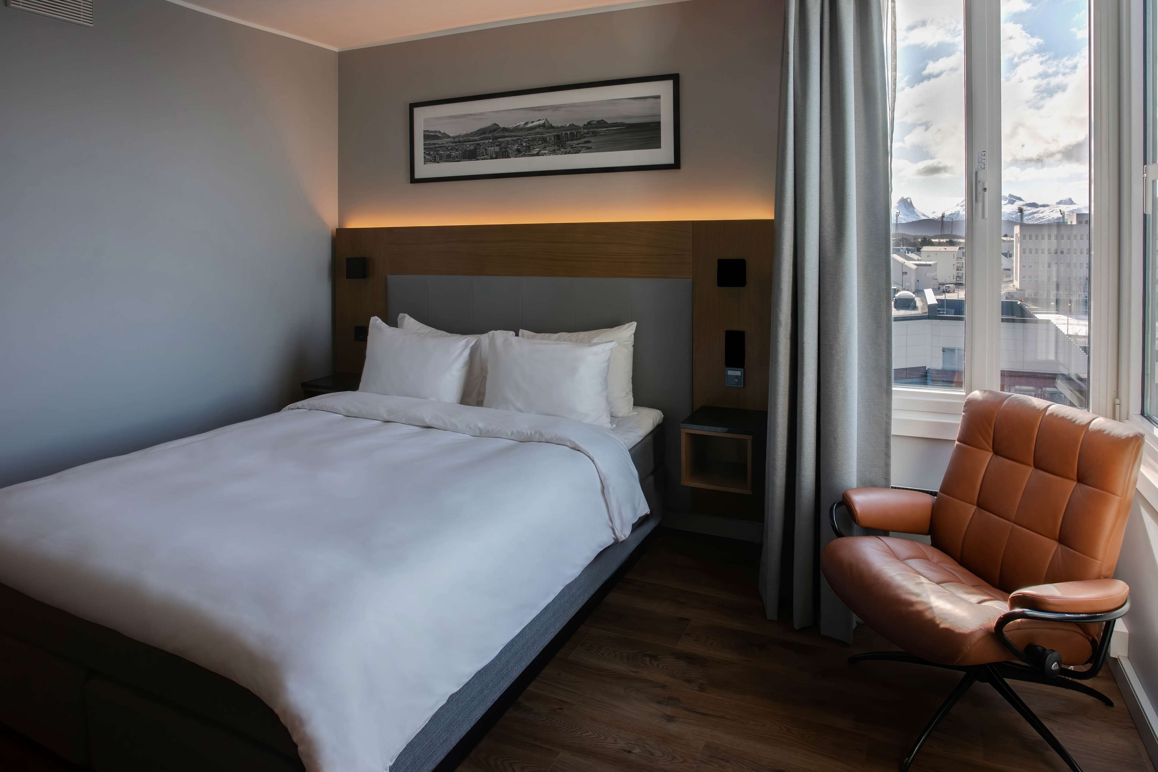 A hotel room with a bed, a chair, a nightstand, a framed picture on the wall, and a window with a view of buildings and mountains.