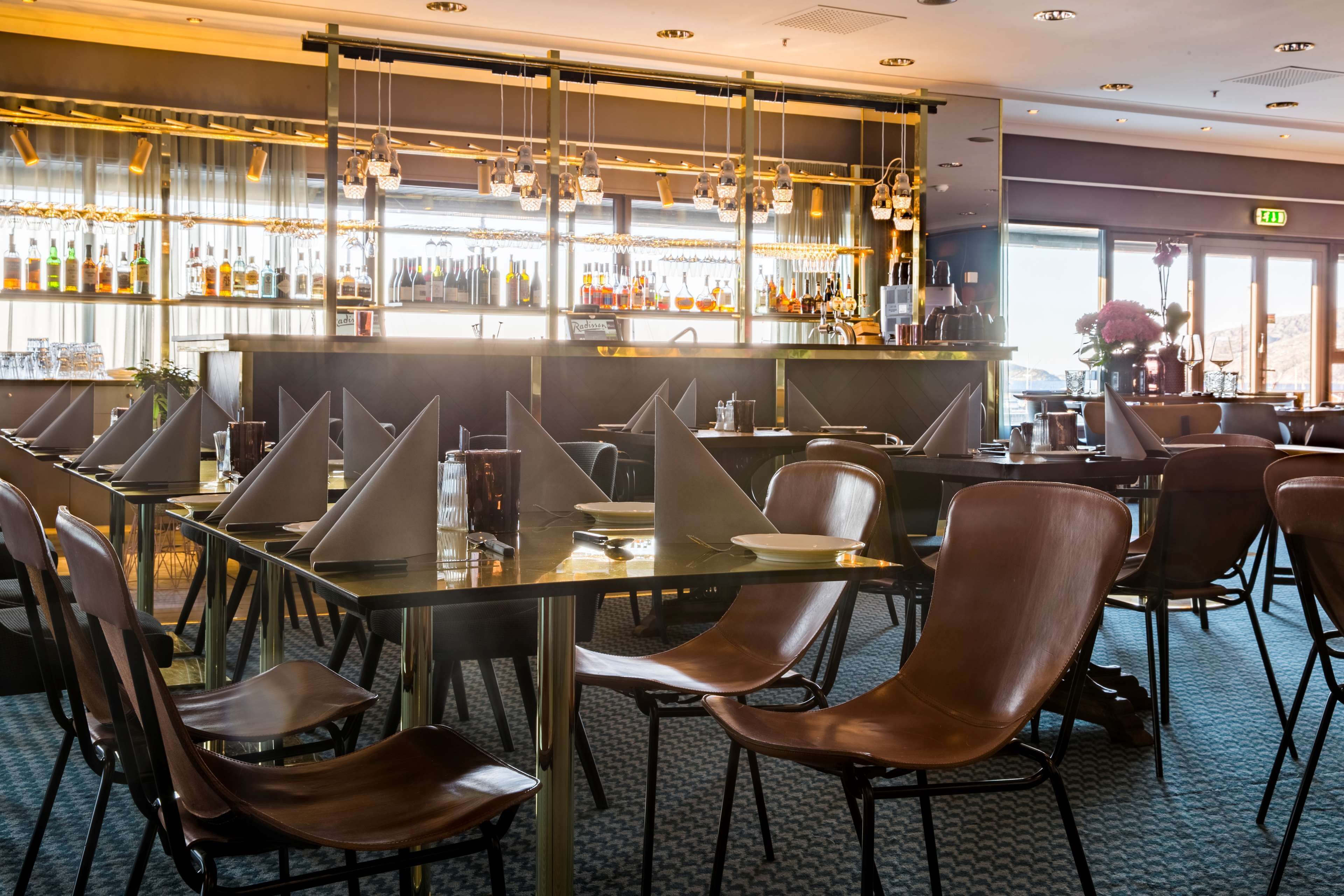 Un espace bar doté de nombreuses tables et chaises, d’un comptoir bien garni et offrant une vue sur l’extérieur grâce à de grandes fenêtres.