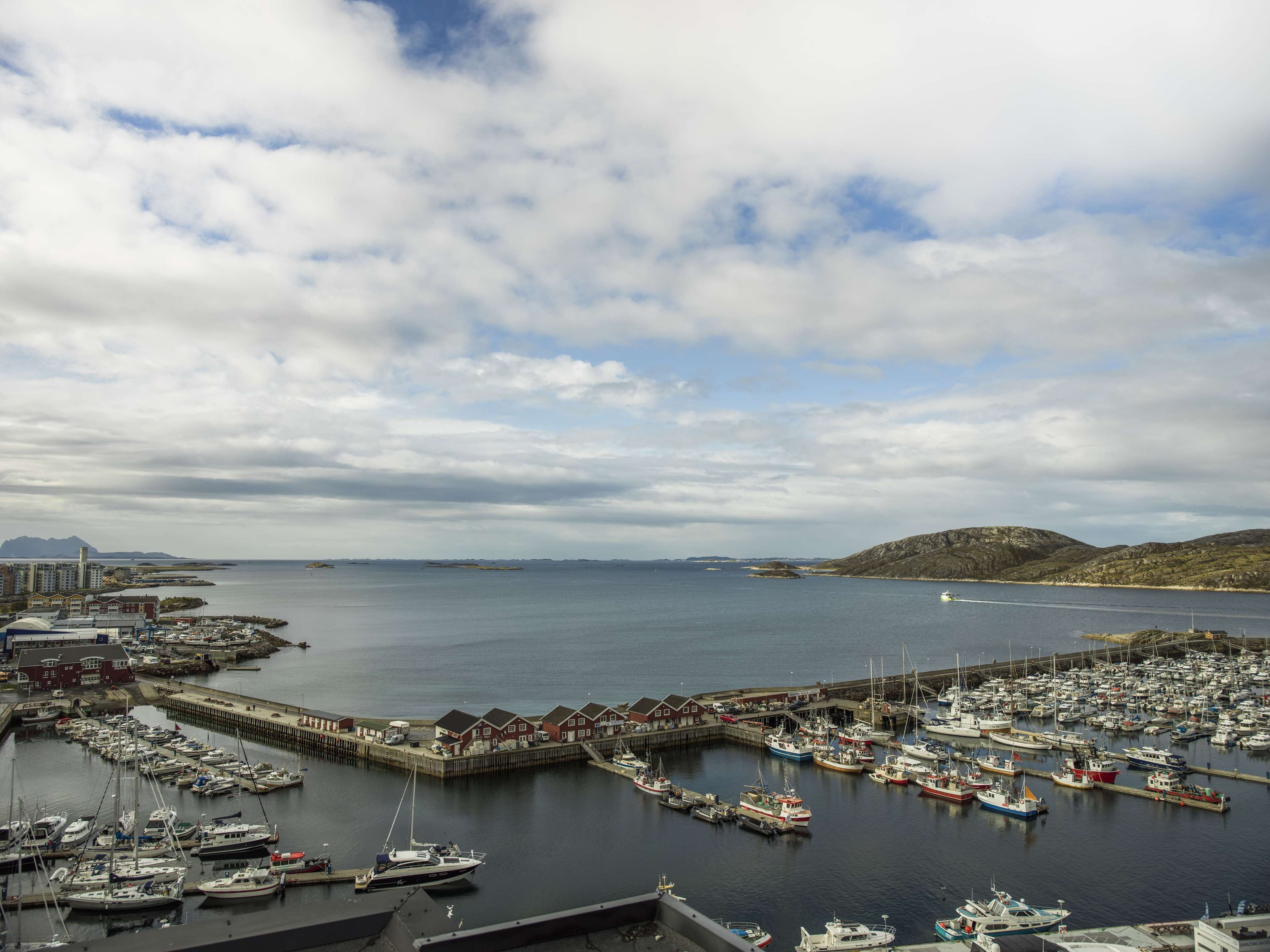 A marina with numerous boats docked, a pier, and a backdrop of hills and a partly cloudy sky.