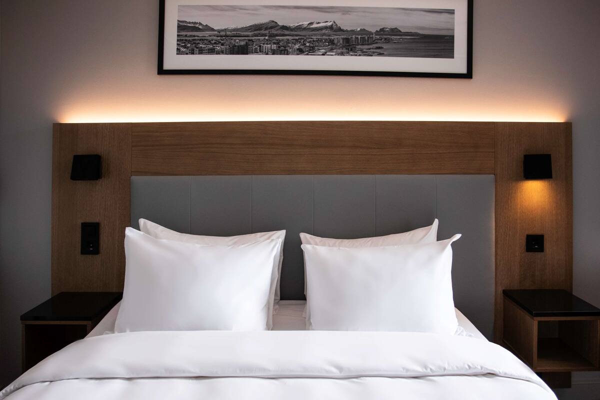 Una cama bien hecha con sábanas blancas, un cabecero de madera y dos mesitas de noche con lámparas negras.
