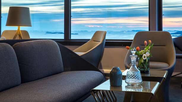 A modern room with a sofa, chairs, a table, a lamp, and a view of the ocean at sunset.