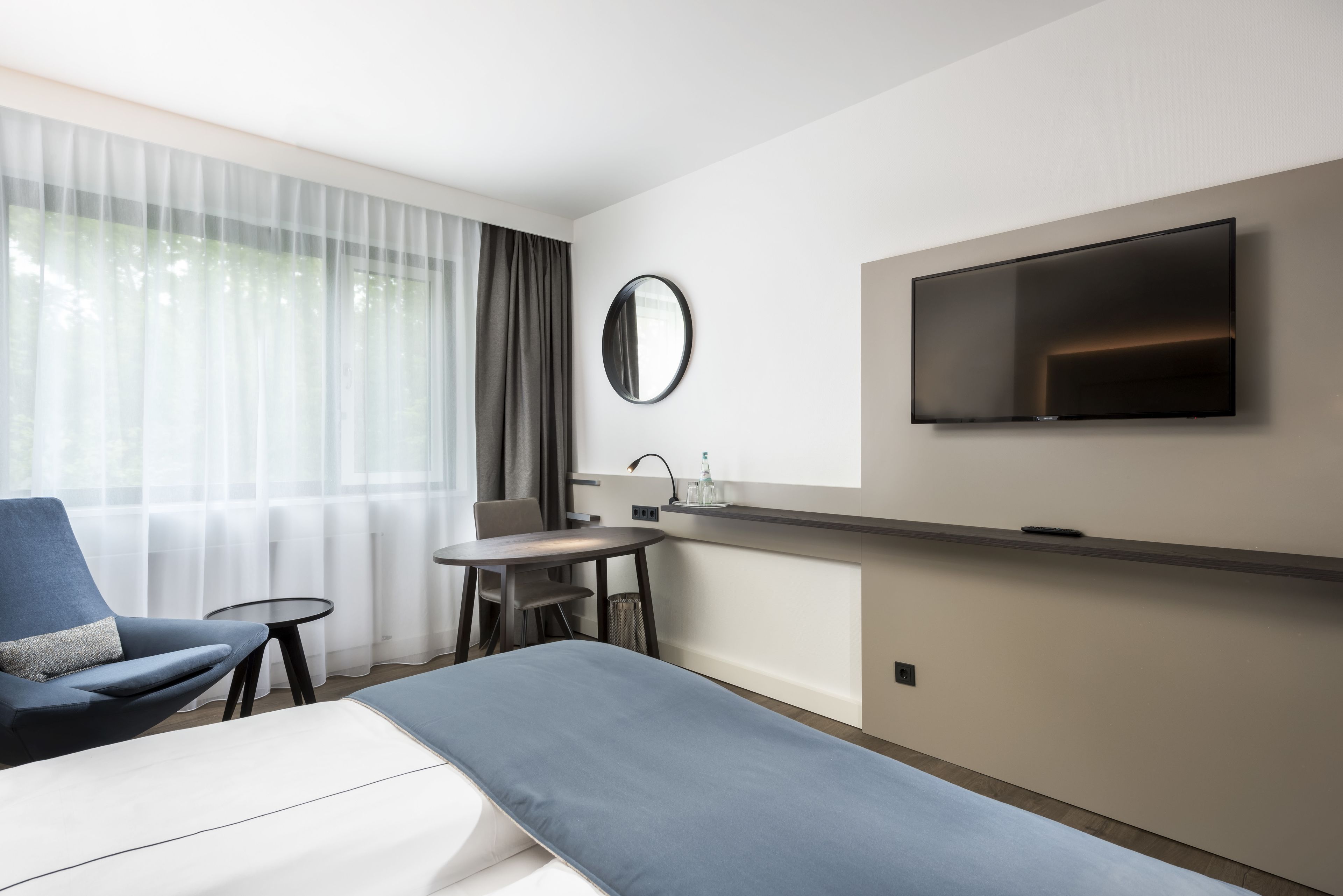 A modern hotel room with a bed, a desk, a chair, a round mirror, and a TV mounted on the wall.