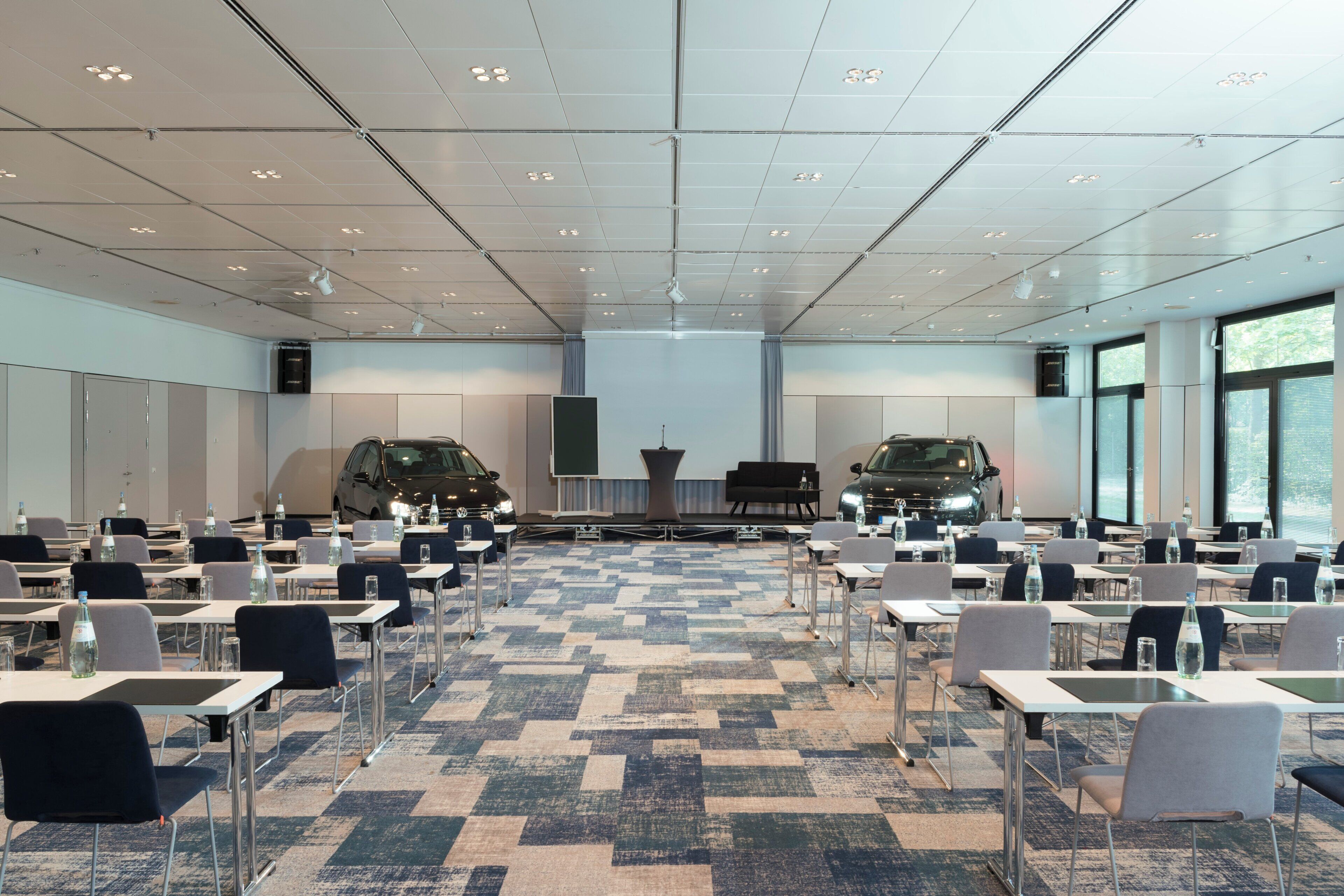 Un salón de conferencias con mesas, sillas, un escenario y dos autos estacionados afuera.