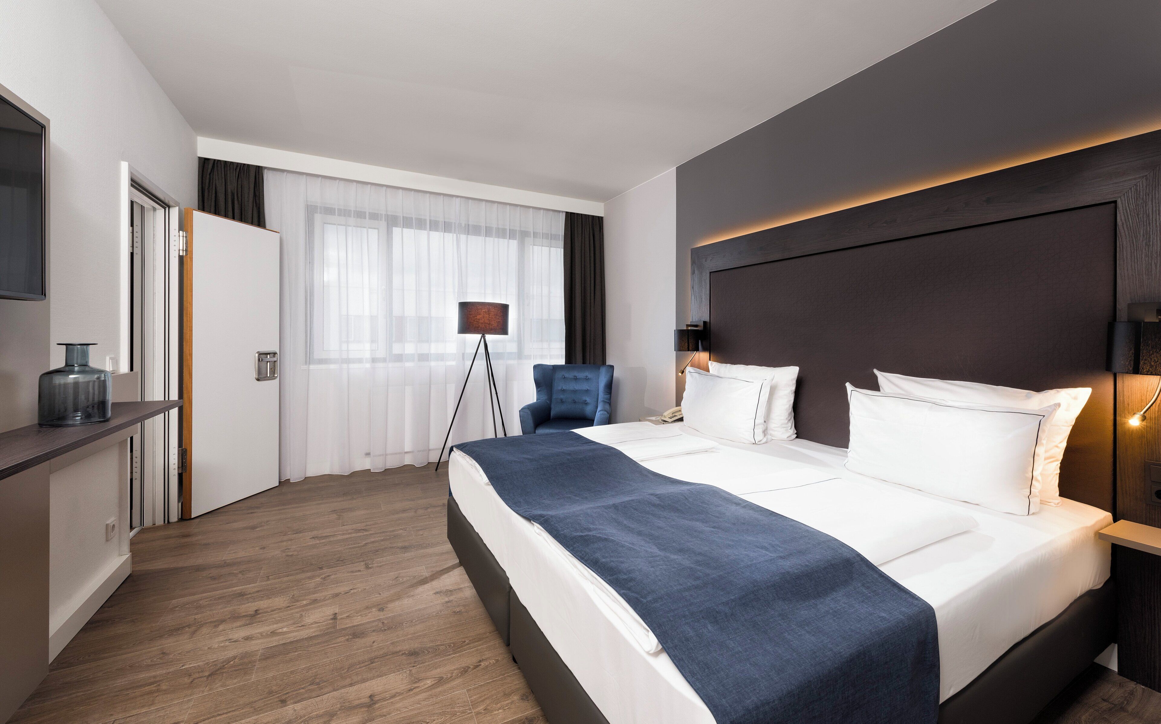 A hotel room with a bed, bedside table, lamp, and a view of the outside.