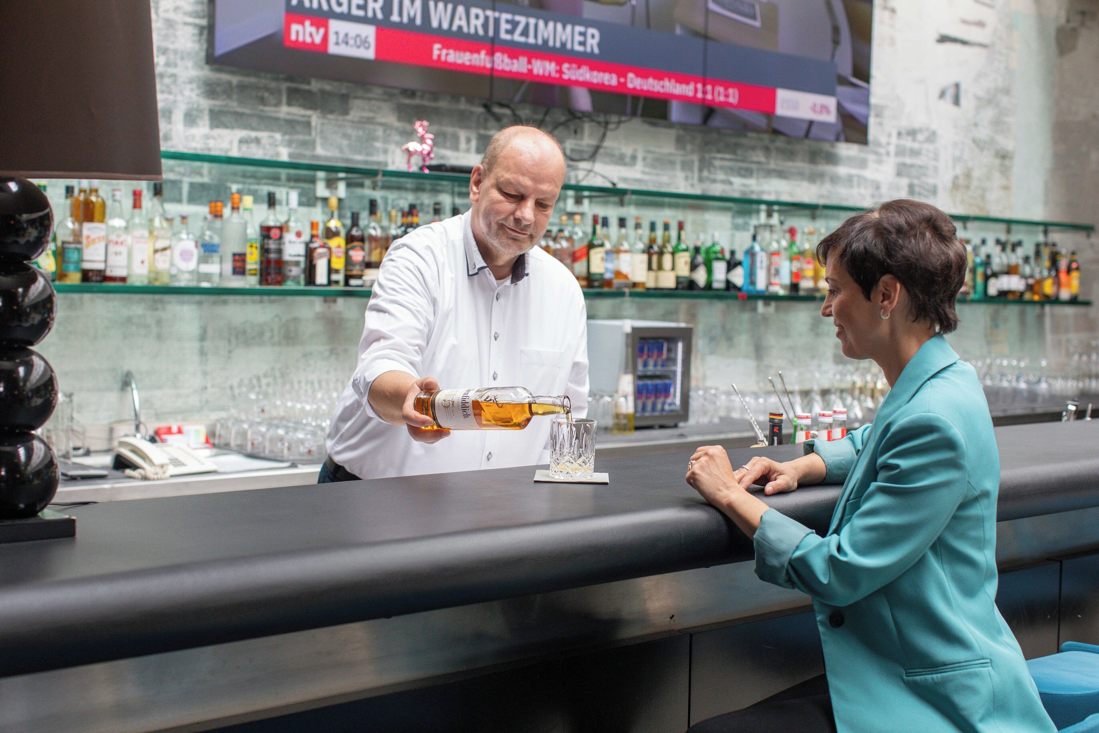 Eine Bar mit einem Barkeeper, der ein Getränk einschenkt, Regale mit verschiedenen Flaschen und einem Schild im Hintergrund.