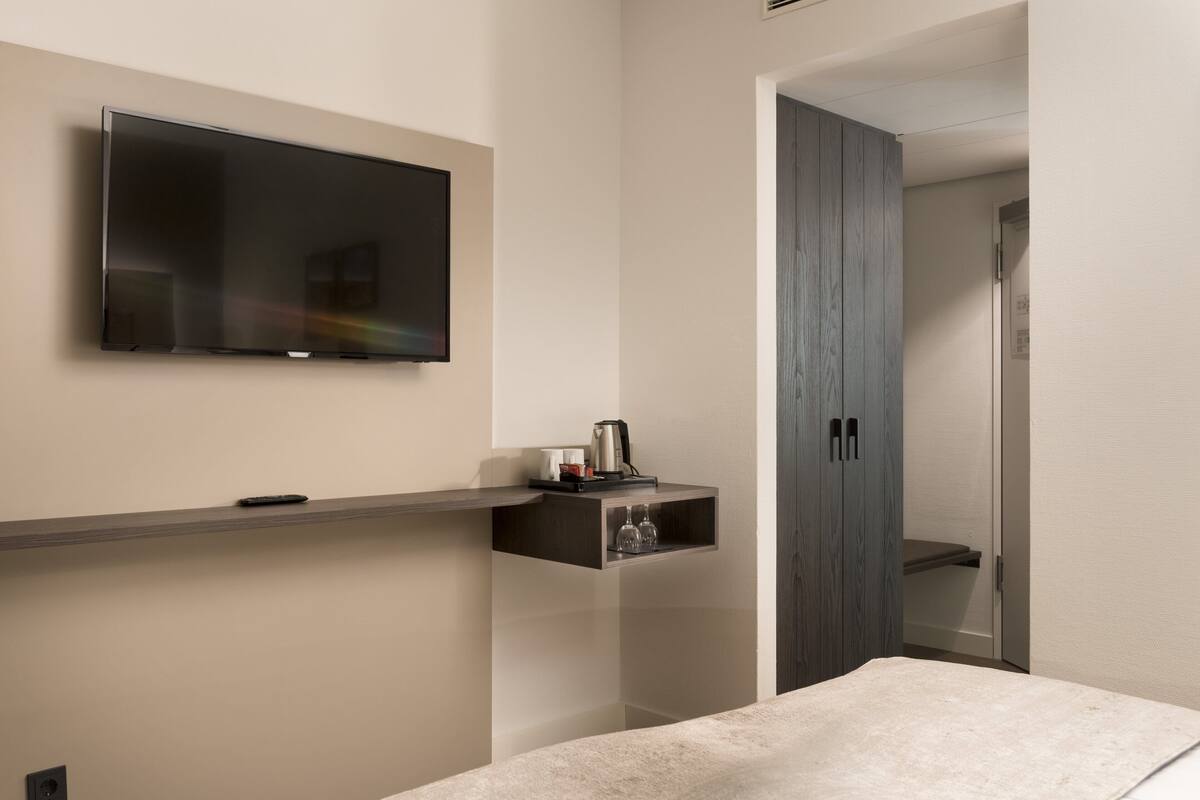 Habitación de hotel moderna con una televisión de pantalla plana montada en la pared, un pequeño estante con una cafetera y vasos, un armario de madera y una cama con cubierta color beige.