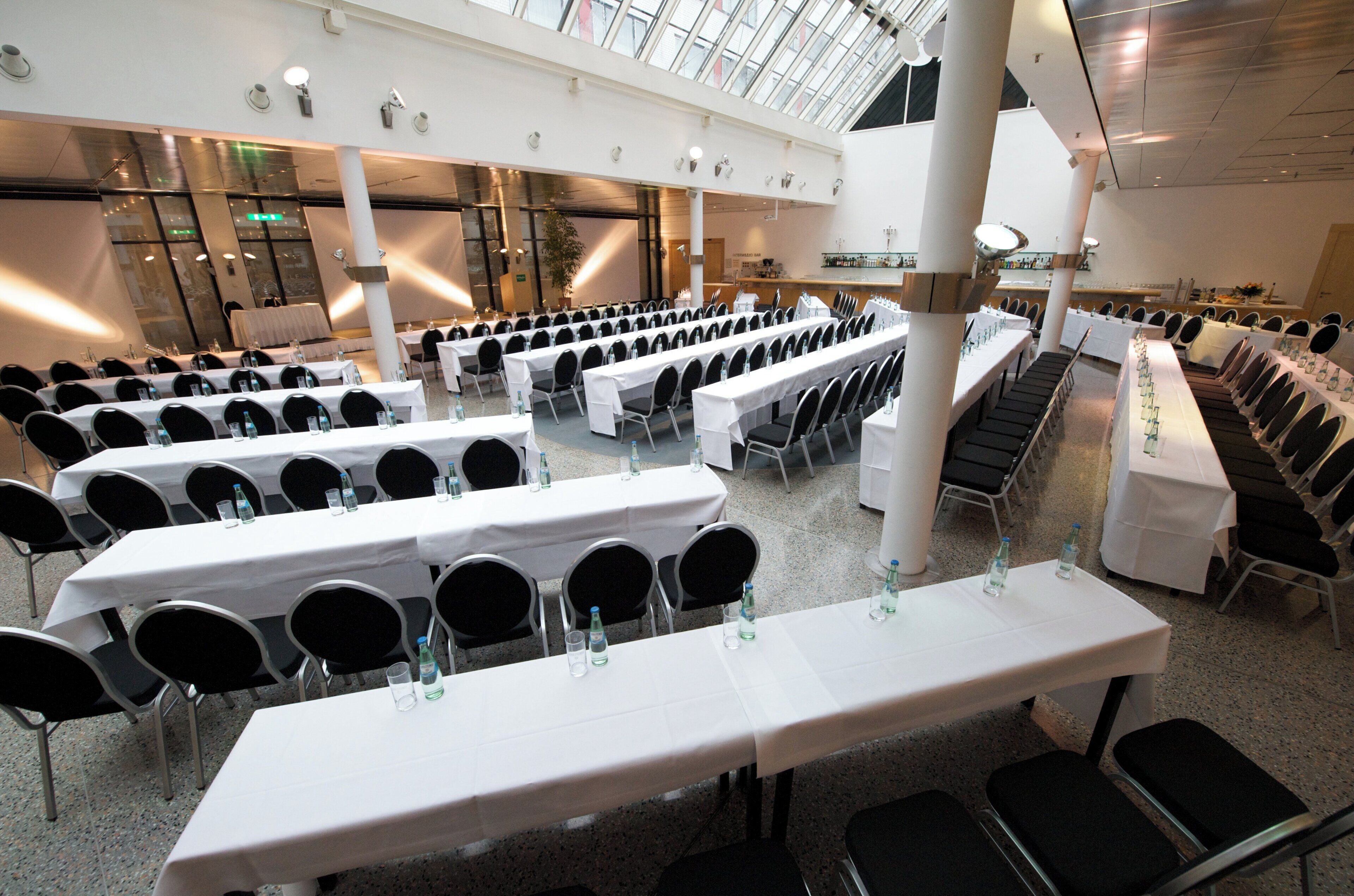 A spacious indoor venue with multiple rows of tables and chairs, a high ceiling with skylights, and a bar area.