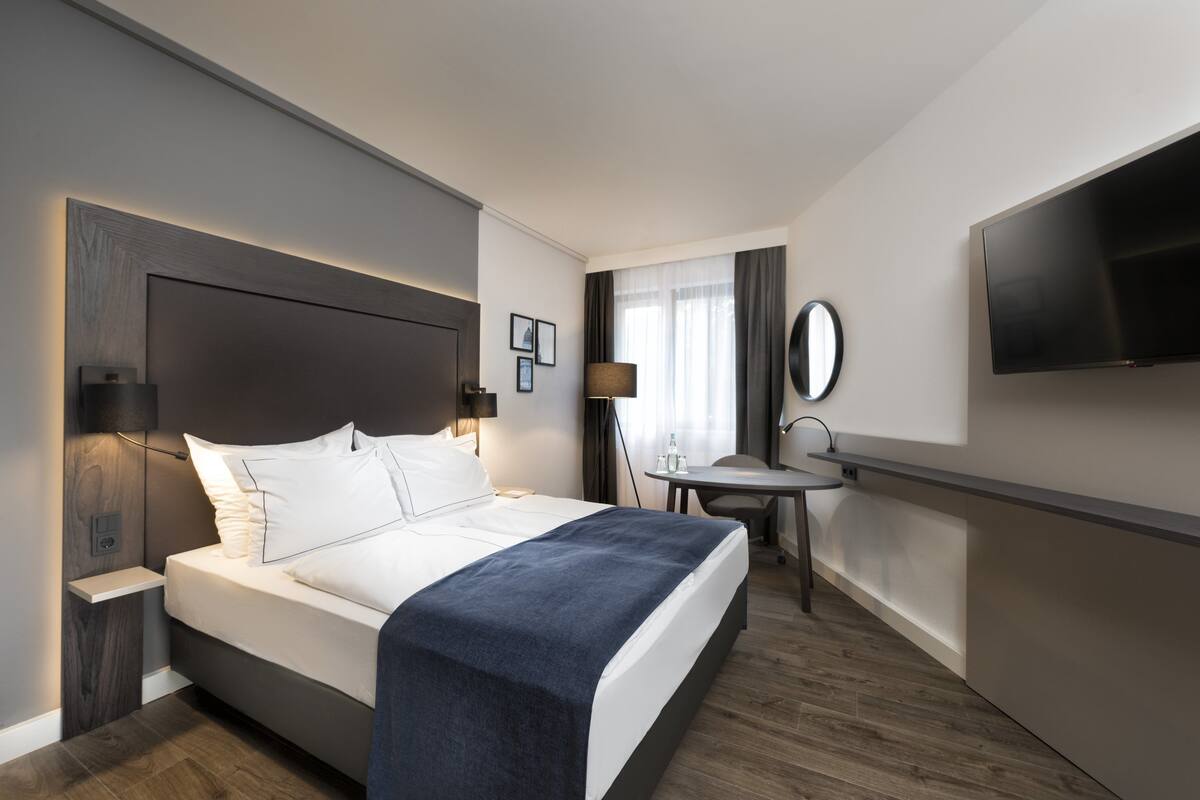 Una habitación de hotel moderna con una cama grande, mesita de noche, lámpara y un televisor de pantalla plana montado en la pared.