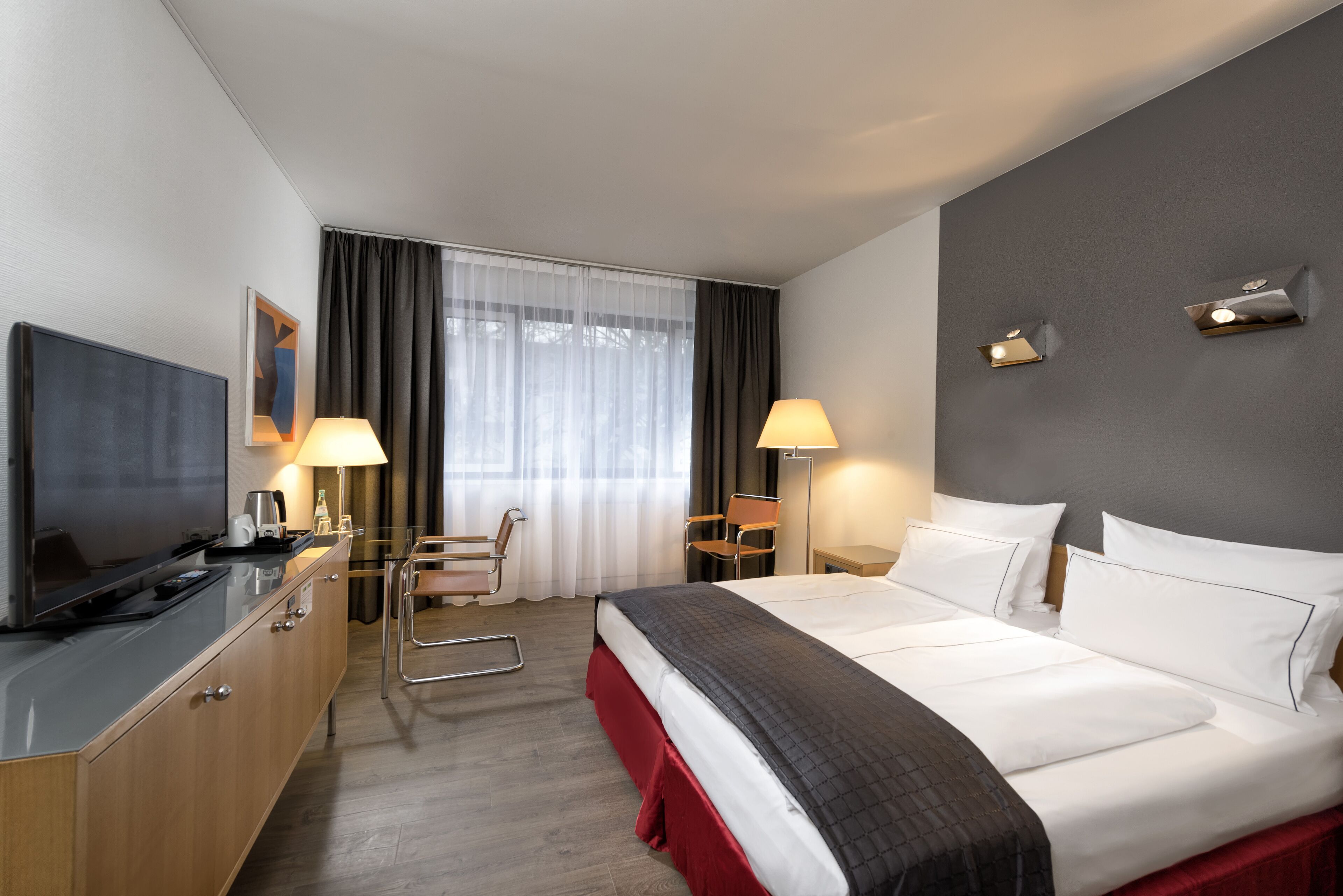 Habitación de hotel con cama, escritorio con televisor, una silla, lámpara y ventana con cortinas.