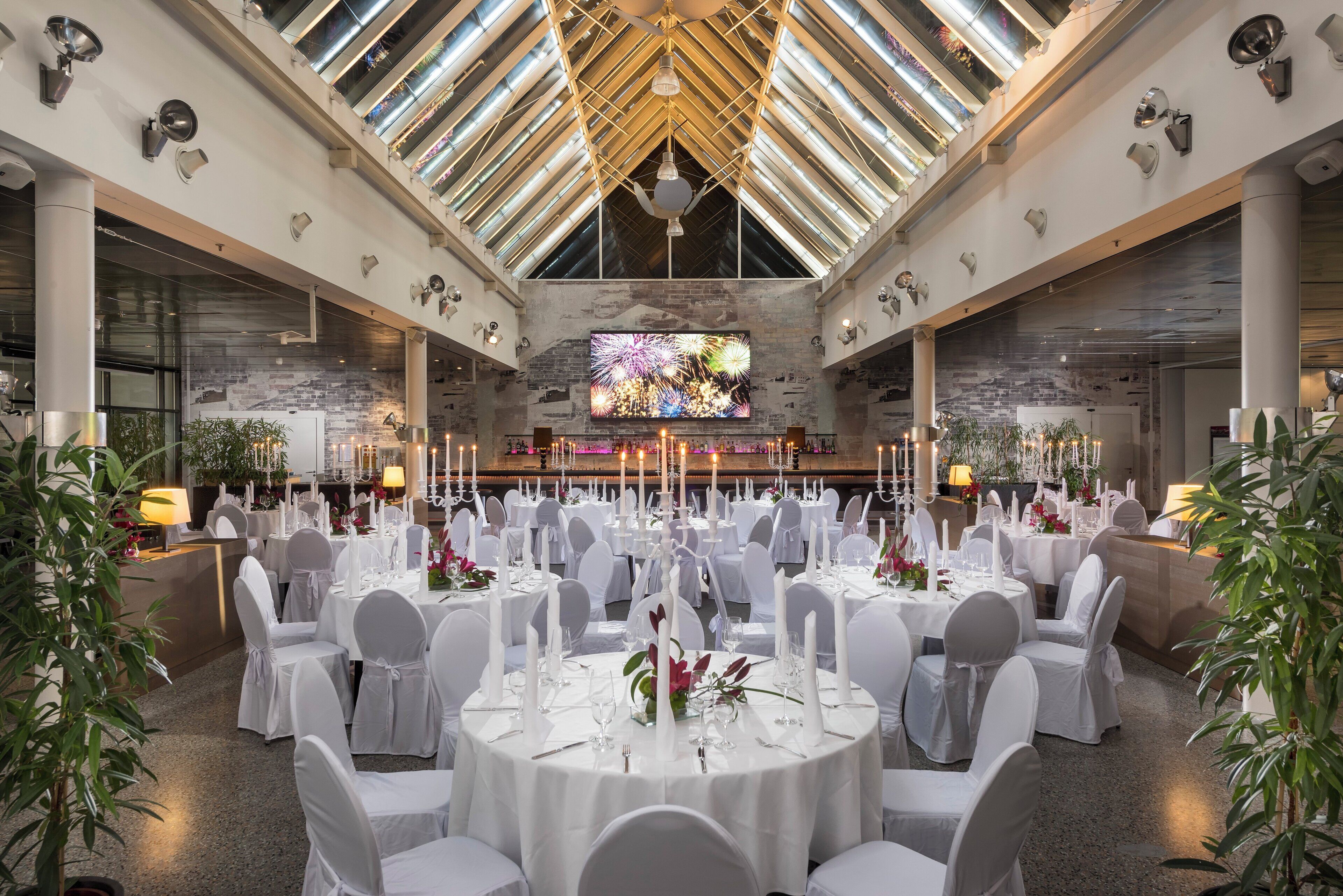 A spacious banquet hall with high ceilings, large windows, and a central stage. Tables are set with white tablecloths and chairs, and the room is decorated with plants and candles.