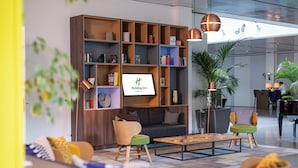 A modern hotel lobby with a large wooden bookshelf, comfortable seating, and a large screen displaying the hotel name. - Holiday Inn Berlin City West by IHG (Berlin)