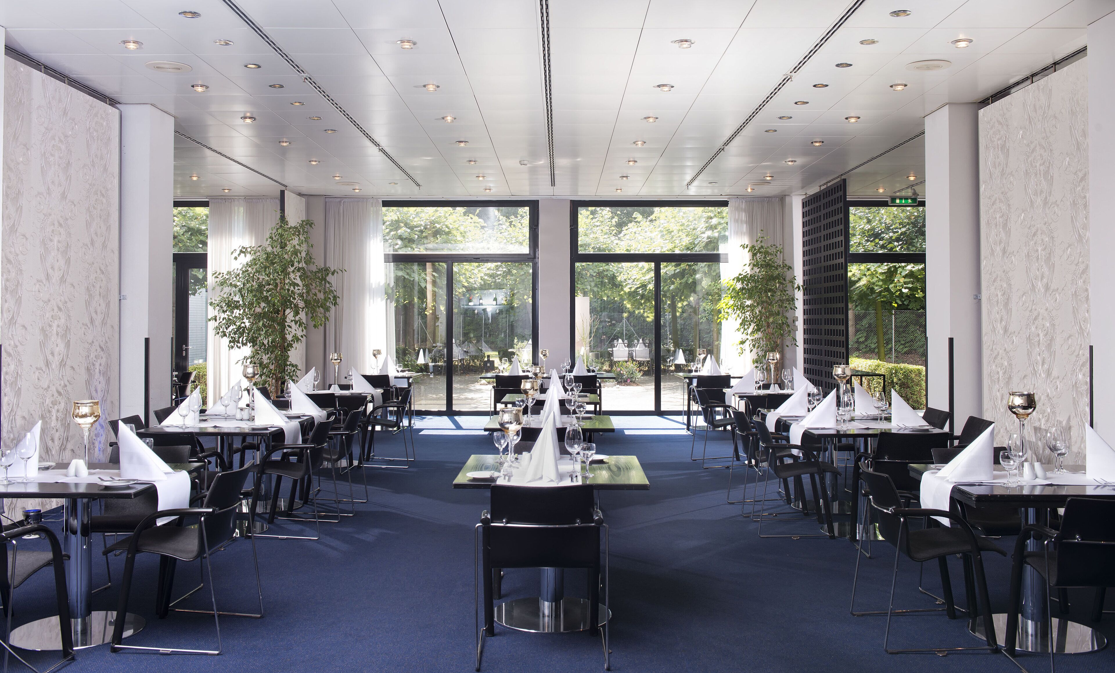 A spacious dining area with multiple tables set for dining, large windows providing natural light, and a view of the outdoors.