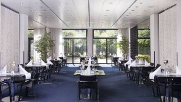 A spacious dining area with multiple tables set for dining, large windows providing natural light, and a view of the outdoors.