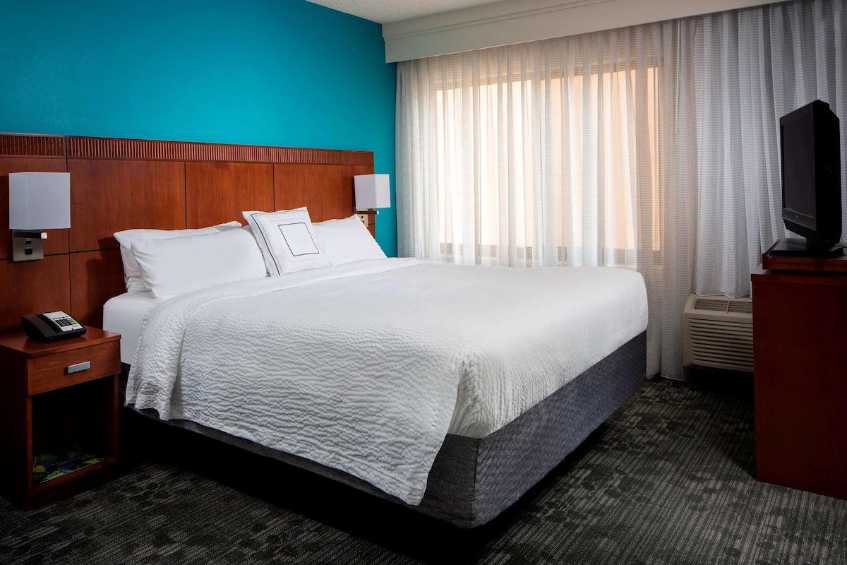 Une chambre d’hôtel avec un grand lit, une tête de lit en bois, du linge de lit blanc, une table de chevet avec un téléphone, un téléviseur posé sur un meuble et une fenêtre avec des rideaux légers.