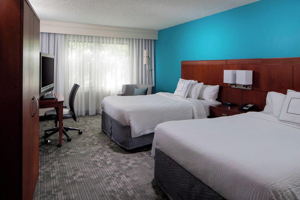 Quarto de hotel com duas camas, uma mesa de trabalho com televisão, uma cadeira e uma janela grande com cortinas.